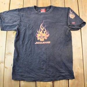 以下が含まれることがあります： 炎の中に頭蓋骨が描かれた黒いTシャツで、"JNCQJEANS"という文字が書かれています。