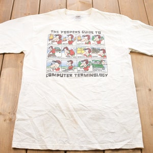 May include: White t-shirt featuring a cartoon illustration of a guide to computer terminology. The text reads "The Yoopers Guide to Computer Terminology".