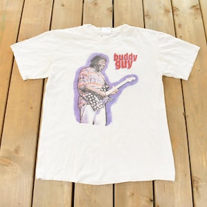 May include: White t-shirt with a graphic of Buddy Guy playing a guitar, and the text "buddy guy" in red. The image is set against a purple background. The shirt is laid flat on a wooden surface.