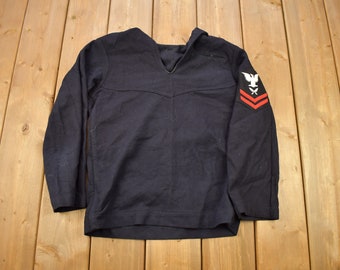 May include: Navy blue vintage sailor shirt with a wide collar and long sleeves. Features a white eagle and red stripes on the left sleeve. The shirt is laid flat on a wooden surface.