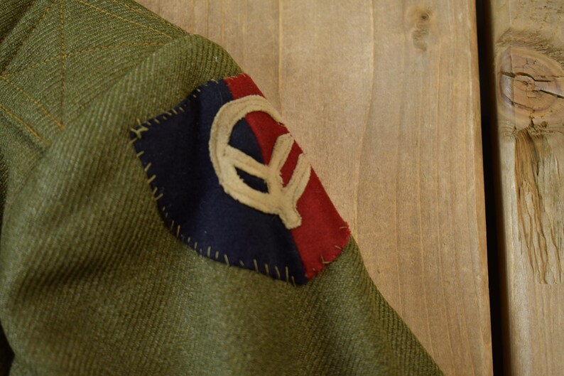 May include: A green military jacket with a red, white, and blue patch sewn on the sleeve. The patch features a stylized symbol.