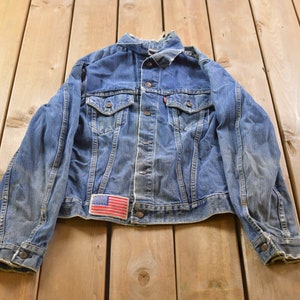 Op de afbeelding: Een blauwe denim jas met een vervaagde look en een klein Amerikaans vlagpatch op de voorkant. De jas heeft een knoopsluiting en twee voorzakken.