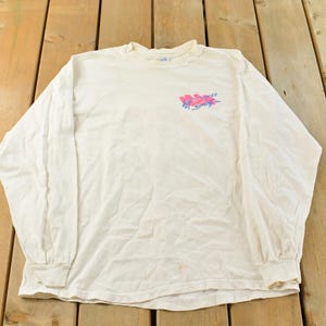 May include: Off-white long-sleeve shirt with a crew neck. The shirt features a colorful graphic on the upper left chest. The graphic appears to be a stylized design with pink and blue hues. The shirt is made of a soft, comfortable material.