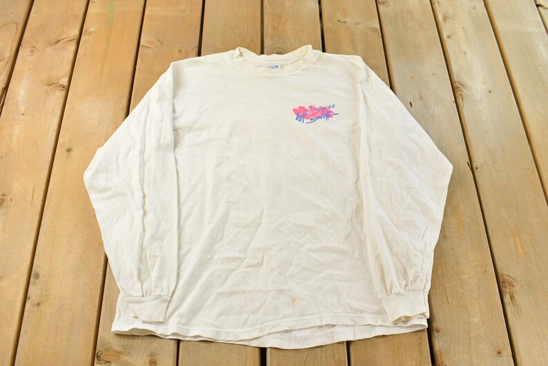 May include: Off-white long-sleeve shirt with a crew neck. The shirt features a colorful graphic on the upper left chest. The graphic appears to be a stylized design with pink and blue hues. The shirt is made of a soft, comfortable material.