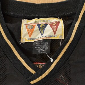 Op de afbeelding: Zwart mesh jersey met een V-hals en gouden strepen langs de kraag en mouwen. De jersey heeft een label met het merk "AVIREX" en de tekst "AUTHENTIC VARSITY SPORTS EQUIPMENT". De maat "XL" staat ook op het label.
