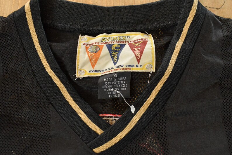 Op de afbeelding: Zwart mesh jersey met een V-hals en gouden strepen langs de kraag en mouwen. De jersey heeft een label met het merk "AVIREX" en de tekst "AUTHENTIC VARSITY SPORTS EQUIPMENT". De maat "XL" staat ook op het label.