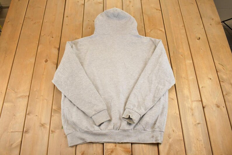 Op de afbeelding: Een lichtgrijze hoodie met een volledige ritssluiting. De hoodie is gemaakt van een zacht, comfortabel materiaal. De capuchon is omhoog en de mouwen zijn lang met geribde manchetten. De hoodie ligt plat op een houten oppervlak.