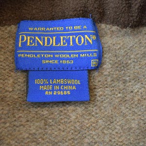 Peut inclure: Gros plan sur une &eacute;tiquette bleue Pendleton avec des lettres dor&eacute;es. On peut lire "WARRANTED TO BE A PENDLETON&reg;" et "PENDLETON WOOLEN MILLS SINCE 1863." En dessous, une petite &eacute;tiquette indique "100% LAMBSWOOL MADE IN CHINA RN 29685." Le fond est un tissu marron textur&eacute;.