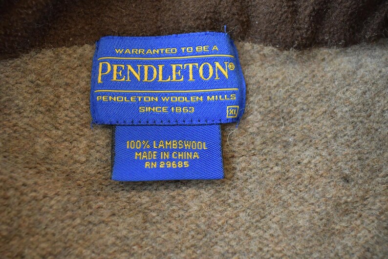 Peut inclure: Gros plan sur une &eacute;tiquette bleue Pendleton avec des lettres dor&eacute;es. On peut lire "WARRANTED TO BE A PENDLETON&reg;" et "PENDLETON WOOLEN MILLS SINCE 1863." En dessous, une petite &eacute;tiquette indique "100% LAMBSWOOL MADE IN CHINA RN 29685." Le fond est un tissu marron textur&eacute;.