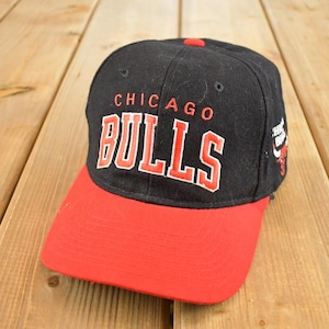 Może przedstawiać: Czarna i czerwona czapka bejsbolowa Chicago Bulls. Czapka ma nazwę drużyny w kolorze białym z czerwonym obrysem z przodu. Daszek jest czerwony, a czapka leży na drewnianej powierzchni.