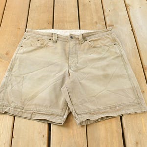 May include: Khaki-colored shorts with a button closure and multiple pockets. The shorts are made of a durable fabric and have visible stitching details. They are laid flat on a wooden surface.