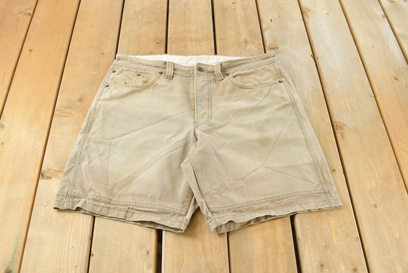 May include: Khaki-colored shorts with a button closure and multiple pockets. The shorts are made of a durable fabric and have visible stitching details. They are laid flat on a wooden surface.