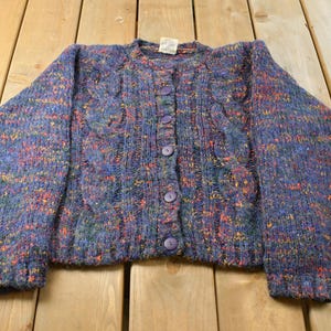 May include: A long-sleeve button-up cardigan sweater in a deep blue hue with a multi-colored flecked pattern. The knit garment features a vertical cable knit design on the front panels, a round neckline, and five matching dark blue buttons.