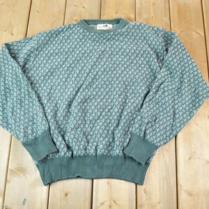 Puede incluir: Un suéter de color verde azulado con un patrón texturizado. El suéter tiene cuello redondo, mangas largas y puños y dobladillo acanalados. La prenda está hecha de un material de punto suave y se exhibe sobre una superficie de madera.