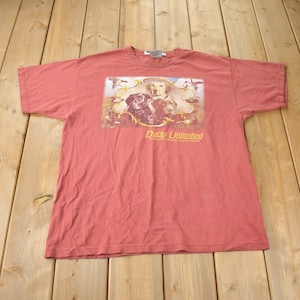 Puede incluir: Una camiseta roja con un gráfico de tres perros y patos. El texto "Ducks Unlimited" está impreso en la camiseta.