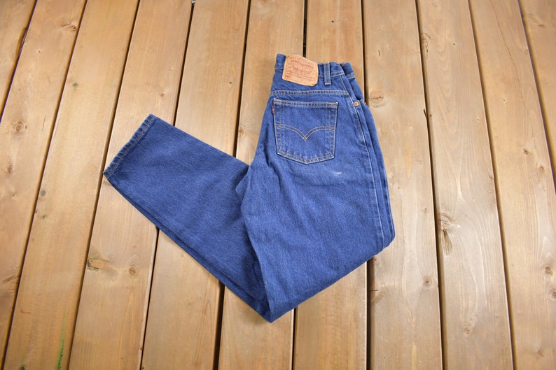 May include: A pair of blue denim jeans with a brown leather patch on the back pocket. The jeans are folded in half and laid out on a wooden surface.