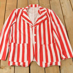 Puede incluir: Blazer a rayas rojas y blancas con dos bolsillos delanteros y tres botones blancos. La chaqueta está tendida sobre una superficie de madera. El blazer tiene mangas largas y solapas con muescas.