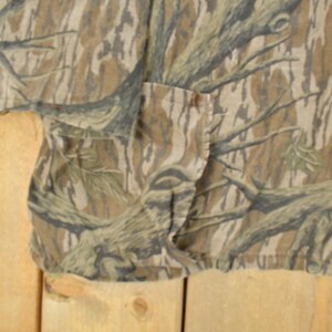 Op de afbeelding: Een shirt met korte mouwen met een camouflagepatroon in bruin, beige en groen. De stof heeft een gedetailleerde print van boomtakken en boomschors. Het shirt heeft een zak en een knoopsluiting.