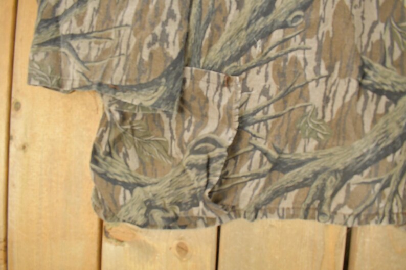 Op de afbeelding: Een shirt met korte mouwen met een camouflagepatroon in bruin, beige en groen. De stof heeft een gedetailleerde print van boomtakken en boomschors. Het shirt heeft een zak en een knoopsluiting.