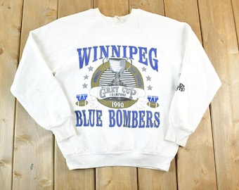 May include: White sweatshirt featuring the text "WINNIPEG BLUE BOMBERS" in blue, with a Grey Cup Champions 1990 graphic. The sweatshirt has a crew neck and long sleeves. The design includes a trophy and stars.