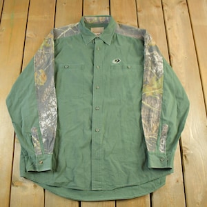 May include: Green button-down shirt with camouflage patterned sleeves and shoulder accents. The shirt has two chest pockets and a small logo on the chest. The long-sleeved shirt is made of a durable fabric.