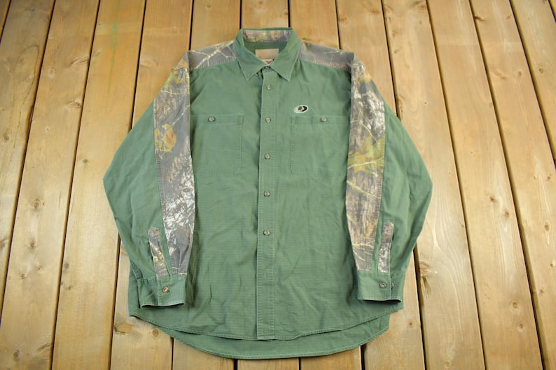 May include: Green button-down shirt with camouflage patterned sleeves and shoulder accents. The shirt has two chest pockets and a small logo on the chest. The long-sleeved shirt is made of a durable fabric.