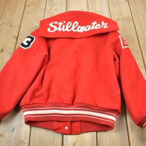 Könnte beinhalten: Rote Varsity-Jacke mit weißen und schwarzen Streifen an Ärmeln und Bund. Der Kragen ist hochgeschlagen und trägt den Schriftzug "Stillwater" in weiß. Die Jacke hat eine weiße Nummer "3" am linken Ärmel.