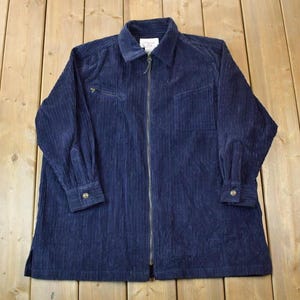 Könnte beinhalten: Eine dunkelblaue Cordjacke mit Reißverschluss und zwei Taschen. Die Jacke hat eine gerippte Struktur und einen Kragen.