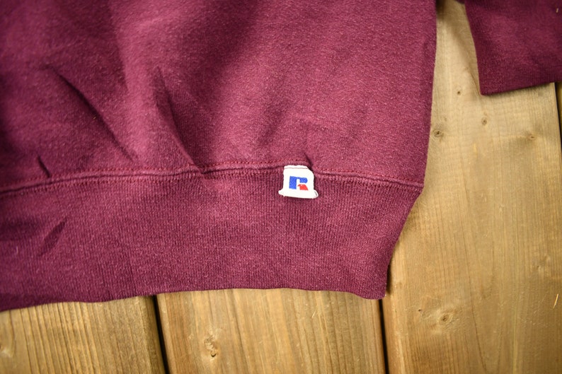 May include: Burgundy sweatshirt with a small blue and white "R" logo tag sewn on the bottom right side.