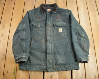 Puede incluir: Una chaqueta de trabajo Carhartt azul oscuro con cuello de pana y un parche con el logotipo de Carhartt en el pecho izquierdo. La chaqueta tiene dos bolsillos grandes en la parte delantera y dos bolsillos más pequeños en el pecho. La chaqueta está usada y tiene un aspecto descolorido.