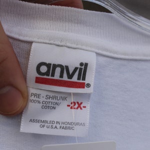 May include: White t-shirt tag with black text that reads "anvil" and "PRE-SHRUNK 100% COTTON/COTON" and "ASSEMBLED IN HONDURAS OF U.S.A. FABRIC". The tag also has a red stripe with black text that reads "-2X-".