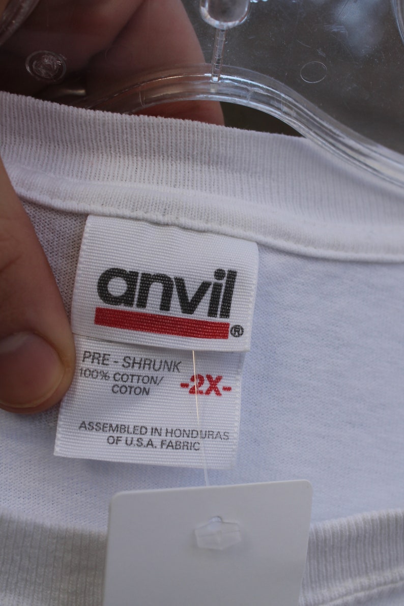 May include: White t-shirt tag with black text that reads "anvil" and "PRE-SHRUNK 100% COTTON/COTON" and "ASSEMBLED IN HONDURAS OF U.S.A. FABRIC". The tag also has a red stripe with black text that reads "-2X-".