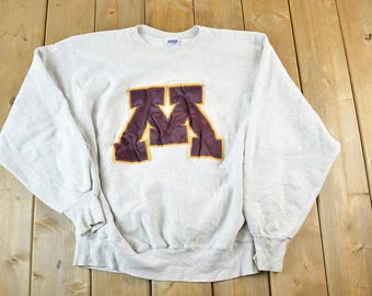 May include: Light gray sweatshirt with a large maroon and gold "M" logo on the front. The long-sleeved crew neck top is made of a soft material and is suitable for casual wear. The sweatshirt is a vintage item.