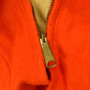K&ouml;nnte beinhalten: Nahaufnahme eines Rei&szlig;verschlusses an einem leuchtend orangefarbenen Kleidungsst&uuml;ck. Der Rei&szlig;verschluss ist aus Metall und hat einen Schieber.