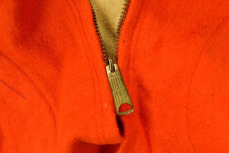 K&ouml;nnte beinhalten: Nahaufnahme eines Rei&szlig;verschlusses an einem leuchtend orangefarbenen Kleidungsst&uuml;ck. Der Rei&szlig;verschluss ist aus Metall und hat einen Schieber.