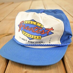 May include: A blue and white baseball cap featuring the text "WORLD'S Greatest GRANDDAD (IT WASN'T EVEN CLOSE.)" in red, blue, and yellow. The cap has a blue peak and a white front panel with the text.