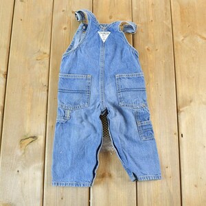 May include: Light blue denim overalls with multiple pockets and a white tag. The overalls are designed for a child and are laid flat on a wooden surface. The overalls have a classic, vintage style.