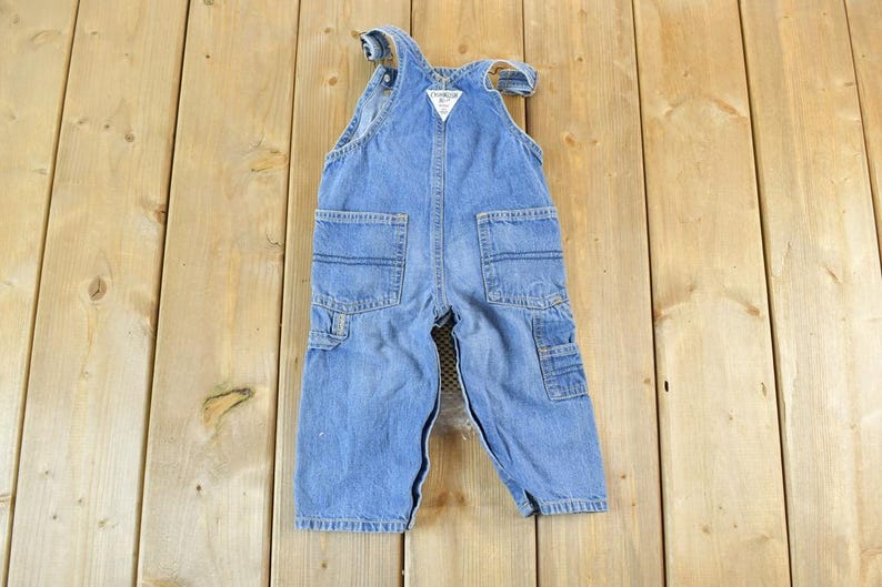 May include: Light blue denim overalls with multiple pockets and a white tag. The overalls are designed for a child and are laid flat on a wooden surface. The overalls have a classic, vintage style.