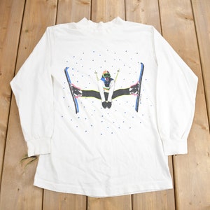 May include: White long-sleeved shirt featuring a graphic of a person skiing in a snowy scene. The graphic is in bright colours, including blue, red, yellow, and green.