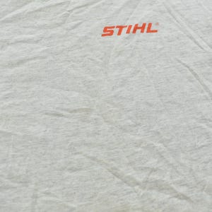 Op de afbeelding: Een lichtgrijs gem&ecirc;leerd T-shirt met het oranje STIHL-logo op de bovenkant van de borst. De stof heeft een zichtbare textuur en vertoont natuurlijke plooien en kreukels over het gehele oppervlak van het kledingstuk.
