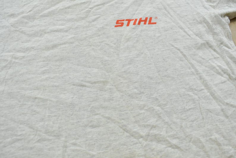 Op de afbeelding: Een lichtgrijs gem&ecirc;leerd T-shirt met het oranje STIHL-logo op de bovenkant van de borst. De stof heeft een zichtbare textuur en vertoont natuurlijke plooien en kreukels over het gehele oppervlak van het kledingstuk.