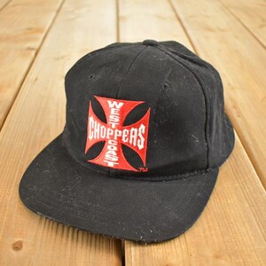 Può includere: Cappellino da baseball nero con visiera piatta. Il cappellino presenta un logo rosso e bianco con la scritta "WEST COAST CHOPPERS" in un design a croce. Il cappellino è realizzato in un tessuto resistente, possibilmente vintage.