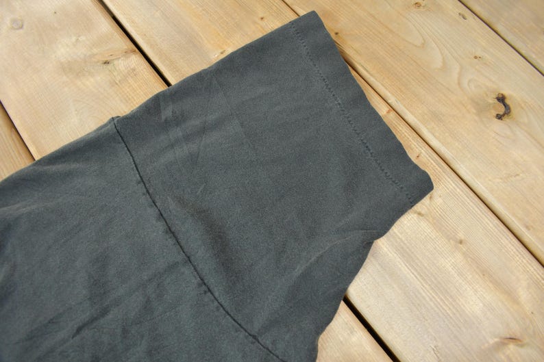 May include: Close-up of the dark gray fabric of a t-shirt. The fabric has a smooth texture and is folded over on itself.