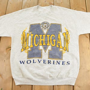 May include: A light gray heathered short-sleeve t-shirt featuring a large navy blue letter M with yellow text reading MICHIGAN and the word WOLVERINES printed below in navy blue. A small circular University of Michigan seal is centered above.