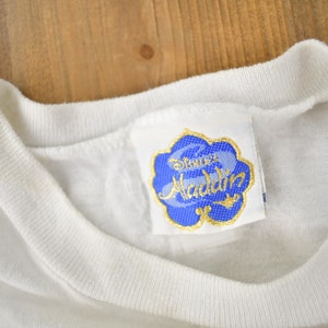 May include: White t-shirt with a blue and gold Disney's Aladdin label attached to the neckline.