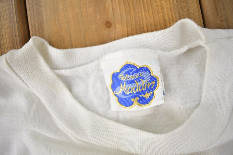May include: White t-shirt with a blue and gold Disney's Aladdin label attached to the neckline.