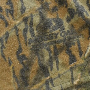 Puede incluir: Primer plano de una tela con estampado de camuflaje en tonos marr&oacute;n, verde y negro. La tela tiene un aspecto texturizado con el texto descolorido "Mossy Oak" visible. El dise&ntilde;o general sugiere un tema de caza o al aire libre.