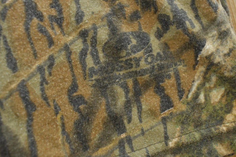 Puede incluir: Primer plano de una tela con estampado de camuflaje en tonos marr&oacute;n, verde y negro. La tela tiene un aspecto texturizado con el texto descolorido "Mossy Oak" visible. El dise&ntilde;o general sugiere un tema de caza o al aire libre.