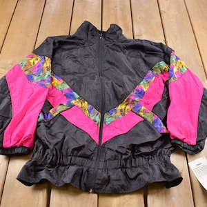 Puede incluir: Chaqueta cortavientos negra con paneles de estampado floral rosa, amarillo y verde. La chaqueta tiene cierre de cremallera y cordón en la cintura.