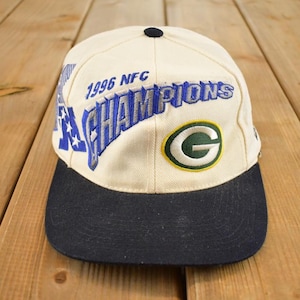 May include: Cream-coloured baseball cap with a navy blue brim. The cap features the text "1996 NFC CHAMPIONS" in blue and white, along with a green and yellow logo. A black button is on top.